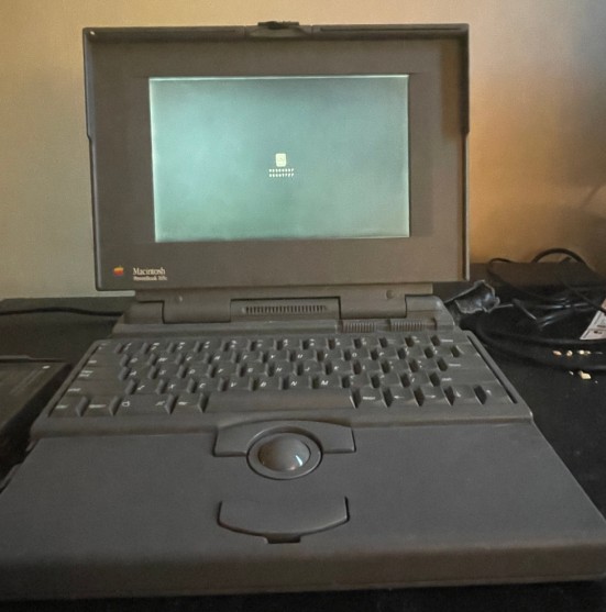 an early example of an apple powerbook. the model is a 165c. it features a color screen, and had an operating system that supported color. the powerbook sits atop a desk with a floppy disk icon on the screen. a question mark appears and disappears within this floppy disk, suggesting no operating system can be found to boot from